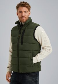 Green padded vest with a high collar, front zipper, two side pockets, and a chest pocket. Worn over a light-colored long-sleeve shirt.