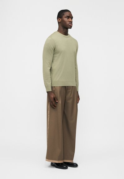 Jeune homme debout, portant un pull à manches longues vert clair, un pantalon ample à jambes larges de couleur marron avec des bandes latérales, et des chaussures noires, sur un fond uni.