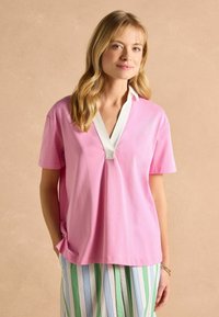 Ružová košeľa s krátkym rukávom a bielym výstrihom do V, s detailom na prednej lége, vyrobená z jemného materiálu. Pod ňou sú viditeľné pruhované nohavice v svetlých farbách.