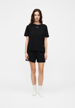 Ung kvinde med langt mørkt hår iført en sort BOSS T-shirt, sorte shorts og hvide sokker, stående mod en ensfarvet hvid baggrund.