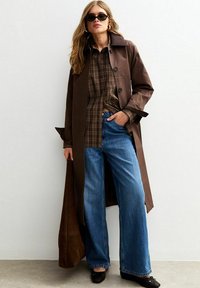 Brown trench coat over a plaid button-up shirt, paired with wide-leg blue jeans. Black sunglasses and shoes complete the look.