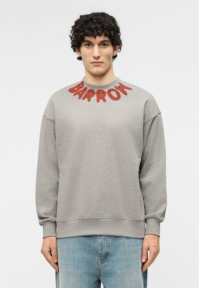 Νεαρός άνδρας με σγουρά μαλλιά, φοράει γκρι φούτερ με κόκκινο κείμενο "BARROW" κοντά στον λαιμό και ανοιχτόχρωμο μπλε τζιν, στέκεται μπροστά σε λευκό φόντο.