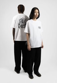 White cotton t-shirts with black graphic designs, one larger on the back, paired with loose black pants and black shoes.