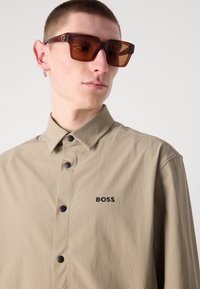 Camicia button-up beige con logo nero, dotata di colletto e tessuto testurizzato. Occhiali da sole quadrati e scuri con lenti di colore marrone.