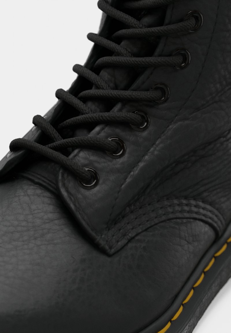 Acercamiento de una bota de cuero negra con cordones negros redondos, ojales de metal y costura amarilla a lo largo del borde de la suela.