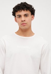 Ilga piedurkne krēmbaltā krekls ar apaļu kakla izgriezumu, centrā ar diskrētu Calvin Klein logotipu, izgatavots no gluda, viegla auduma.