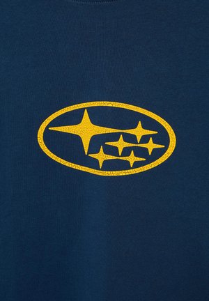 Logo galben crăpat Subaru cu șase stele în interiorul unui oval pe un fundal texturat din pânză albastru închis.