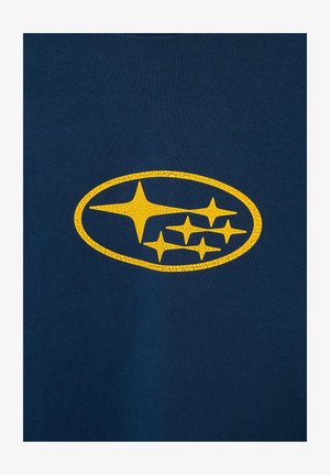 Logo galben crăpat Subaru cu șase stele în interiorul unui oval pe un fundal texturat din pânză albastru închis.