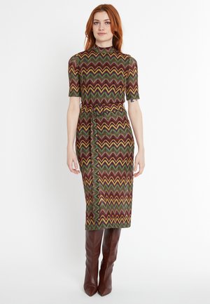 Robe à motifs avec des manches courtes, présentant des motifs en zigzag rouge, vert et jaune. Taille ceinturée et longueur au genou. Accorrdée avec des bottes hautes marron.