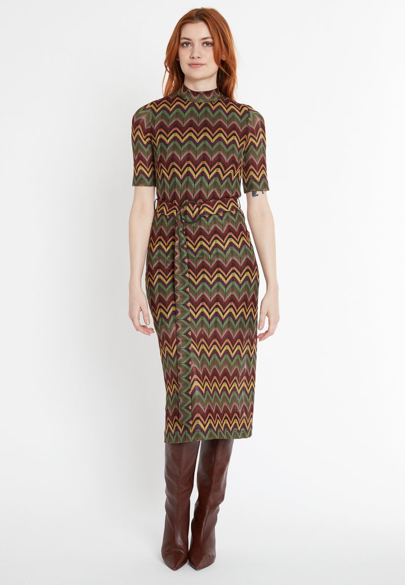 Musterkleid mit kurzen Ärmeln, das Zickzackmuster in Rot, Grün und Gelb aufweist. Mit Gürtel an der Taille und knielang. Trägt braune Stiefel.