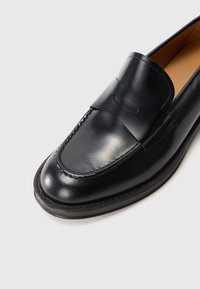 Mocassin en cuir noir avec un bout arrondi, des coutures décoratives et une bande décorative sur le dessus. Texture lisse et design minimaliste.