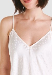 La camiseta blanca sin mangas presenta una suave textura satén, tirantes finos ajustables, un escote en forma de v y un sutil patrón cuadrado con un lazo central como acento.
