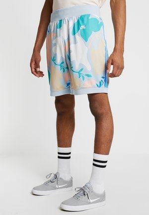 Męskie nogi w pastelowych niebieskich spodenkach Nike w kamuflażu, białe skarpetki z czarnymi paskami oraz szare buty sportowe Nike stojące na białym tle.