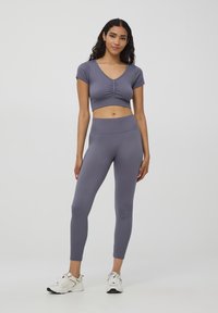 Jasnofioletowy prążkowany crop top z krótkimi rękawami i marszczonym detalem na środku, w komplecie z dopasowanymi legginsami o wysokim stanie. Białe buty sportowe.