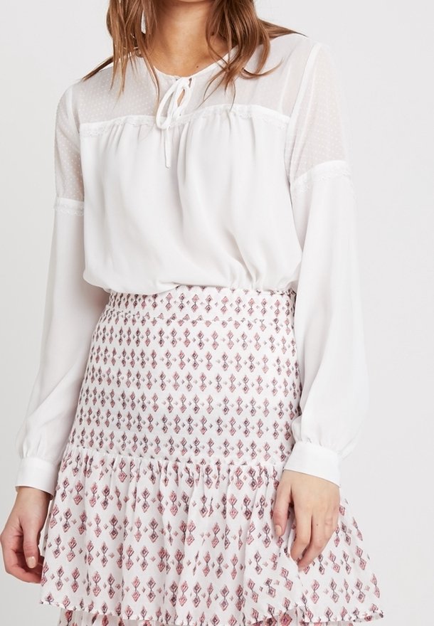 Blusa bianca leggera con dettaglio al collo a fiocco e maniche lunghe trasparenti, abbinata a una gonna a ruota con volant e fantasia caratterizzata da motivi rosa e rossi.