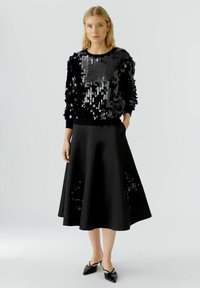Femme portant un haut à manches longues orné de sequins noirs, une jupe midi noire avec des détails floraux et des chaussures à talons pointus noires.