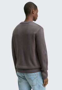 Grauer Strickpullover mit Rundhalsausschnitt, gerippten Bündchen und Saum. Das Material ist strukturiert und wird mit hellblauen Jeans kombiniert.