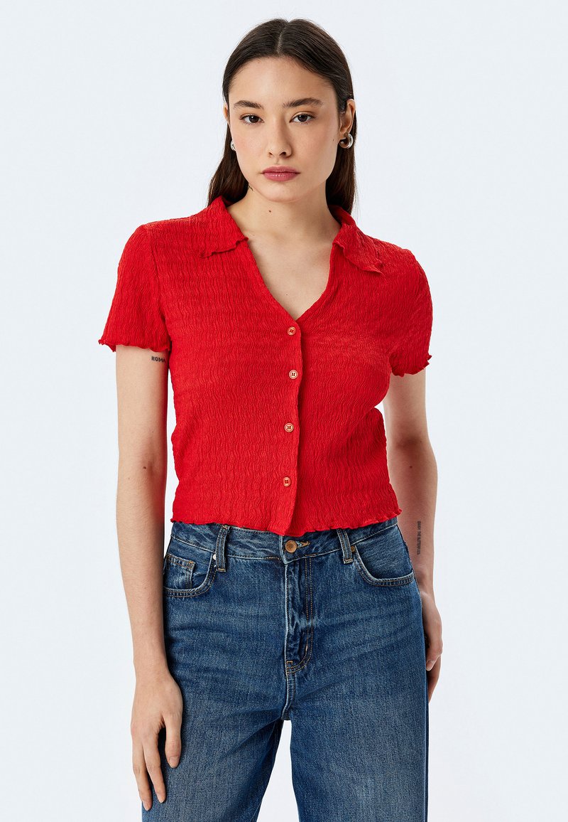 Camicia rossa a maniche corte con colletto, caratterizzata da una texture sfrangiata e piccoli bottoni, abbinata a jeans blu a vita alta.