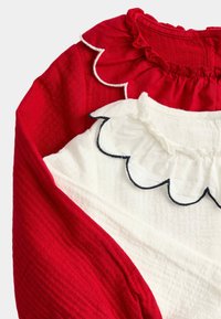 Två skjortor staplade ovanpå varandra, en röd med volangkrage och texturerat tyg, den andra vit med vågad krage och blå kant.