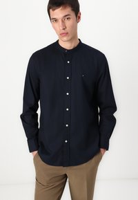 Camicia blu navy con colletto a banda, tessuto leggero e bottoni bianchi. Presenta un piccolo logo ricamato sul petto.