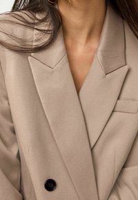 Blazer beige à double boutonnage avec revers crantés. Tissu texturé, doté d'une poche poitrine et d'un détail de bouton noir.