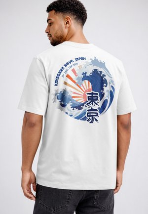 Mann trägt weißes T-Shirt mit großem Motiv einer blauen Ozeanwelle, aufgehender Sonne und japanischem Text auf dem Rücken.