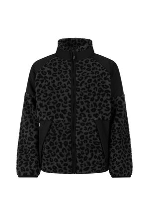 Jong persoon draagt een fleecejas met luipaardprint, beige leggings, zwarte beanie en zwarte snowboots, staand met één hand op de heup.