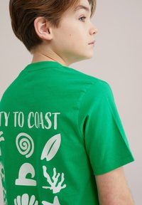 Zielona bawełniana koszulka z białymi nadrukami z motywami morskim oraz napisem "COAST TO COAST" z tyłu.