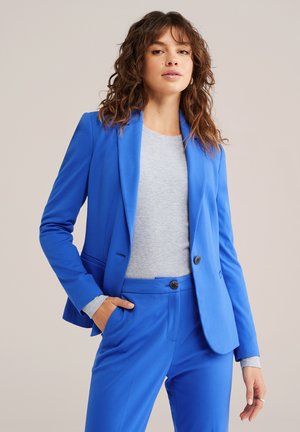 Vrouw met golvend bruin haar, gekleed in een felblauwe blazer en bijpassende broek over een lichtgrijs langemouwen shirt, poserend met één hand in de zak.