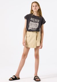 Camiseta gráfica gris con texto blanco, combinada con una falda envolvente beige con bolsillos laterales y sandalias negras con acentos de ribete blanco.