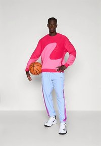 Muž stojí a drží basketbal jednou rukou, na sobě má růžovo-červený svetr, světle modré kalhoty s růžovými pruhy a bílé tenisky.