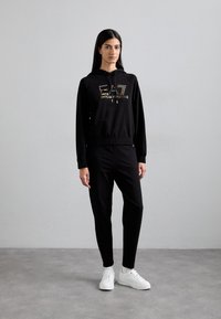 Čierny set s mikinou a nohavicami z mäkkej tkaniny, so zlatým logom na hrudi, elastickými manžetami a voľným strihom, doplnený bielymi teniskami.