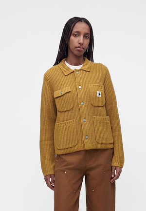 Jeune femme avec de longues tresses portant une veste en maille jaune moutarde avec des poches avant et un pantalon marron sur un fond clair uni.