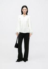 Blouse en satin blanc avec un col en forme de goutte et des manches volumineuses, associée à un pantalon noir ajusté et un petit sac à main noir.
