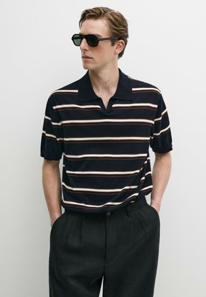 Mann mit schwarzen Sonnenbrillen, kurzärmeliges marineblaues Hemd mit weißen und braunen Streifen und dunkle, hoch taillierte Hose, Hände in den Taschen.