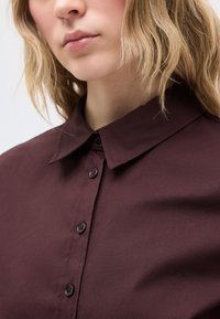 Ξανθό άτομο που φοράει ένα σκούρο καφέ πουκάμισο με κουμπιά και μυτερό γιακά, ορατό από τον λαιμό μέχρι το πάνω μέρος του στήθους.