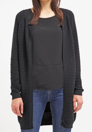 Cardigan nero in maglia con texture a coste, apertura frontale e maniche lunghe, indossato sopra una maglietta nera e jeans blu.