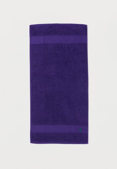 Σκούρα μωβ πετσέτα με δύο οριζόντιες υφασμένες λωρίδες κοντά στις άκρες και ένα μικρό πράσινο κεντητό λογότυπο στην κάτω δεξιά γωνία.