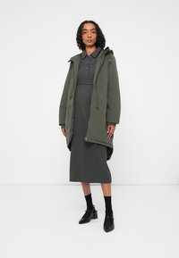 Overdimensioneret grøn parka med snørehætte, lagdelt over en jerseykjole i koksgrå rib. Modellen har sorte sokker og hæle på.