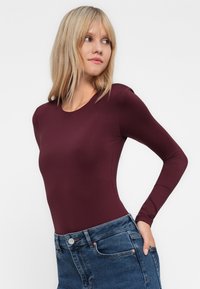 Blond kvinne som har på seg en tettsittende burgunder genser med lange ermer, stukket inn i blå høytlivsjeans, som ser til venstre med hånden i lomma.