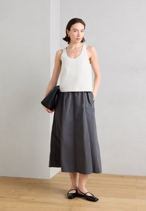 Ženska stoji v notranjosti, oblečena v belo brezrokavnik, sivo midi krilo, črne balerinke in drži črno usnjeno torbico.