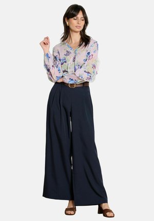 Frau trägt eine rosa Bluse mit Blumenmuster, die in eine hoch taillierte, marineblaue, weit geschnittene Hose gesteckt ist, dazu ein brauner Gürtel und braune Sandalen mit Blockabsatz.