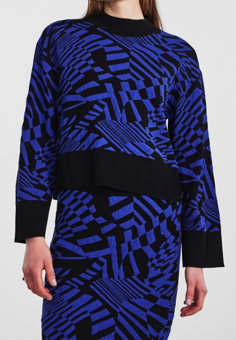 Femme portant un pull bleu et noir à motif géométrique et une jupe assortie avec poignets et encolure côtelés.