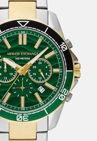 Žalias nerūdijančio plieno laikrodis su auksinėmis detalėmis, tekstūruotas ciferblatas, trys subciferblatai, datos langelis ir tachimetriniai ženklai ant sidabrinės apyrankės.
