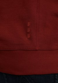 Un primer plano del dobladillo de un sudadera roja, con costuras texturizadas y tres pequeños elementos decorativos rojos en el lado.