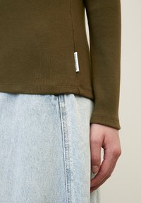 Personne portant un haut à manches longues côtelé olive avec une petite étiquette blanche et un jean en denim bleu clair, la main posée sur la hanche.