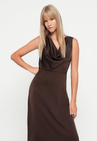 Robe sans manches marron avec un décolleté drapé et une silhouette fluide, fabriquée en tissu doux, présentant un corsage ajusté et une jupe plus longue.