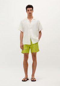 Chemise blanche en lin à manches courtes avec un col boutonné. Short vert vif avec un motif de tête de mort et de tibias bleus. Tongs brunes.