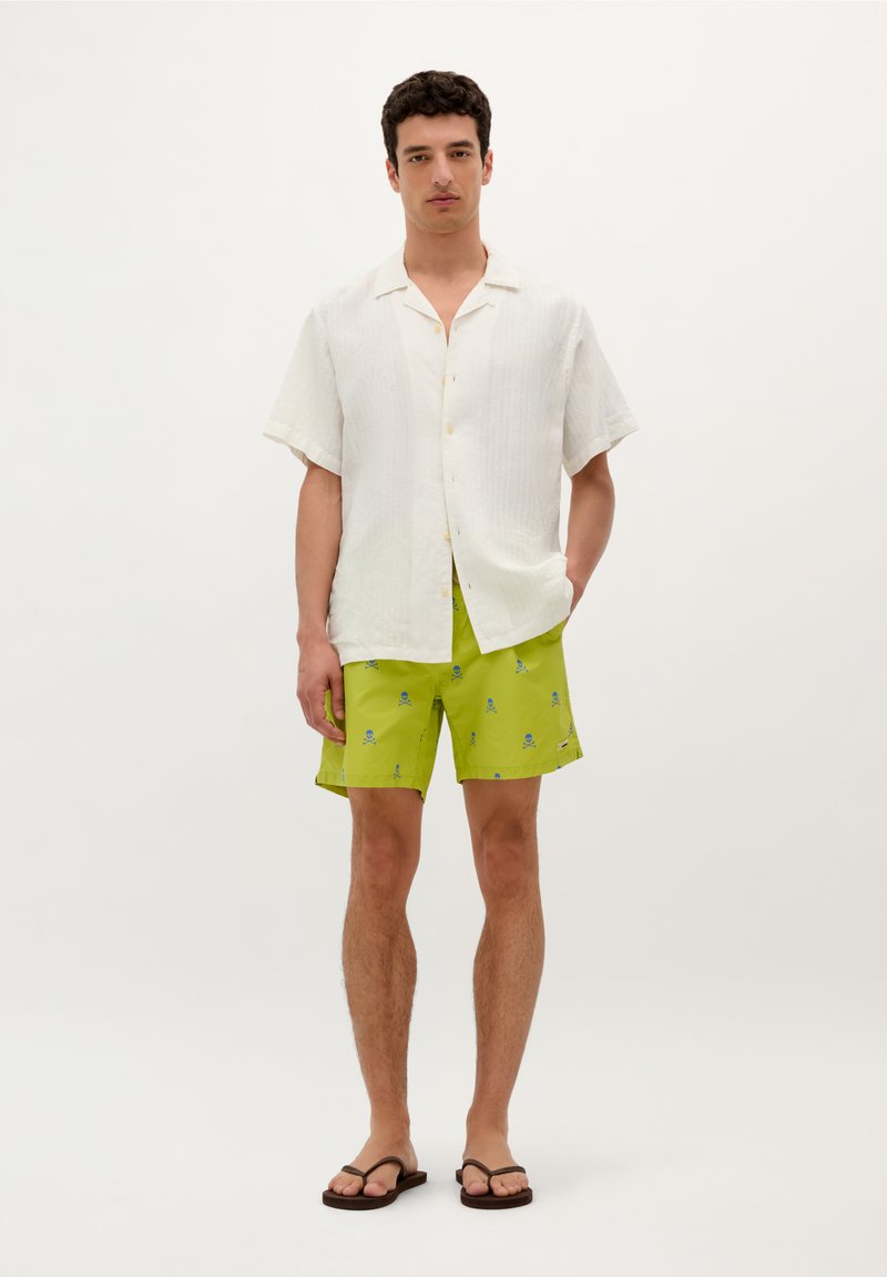 Chemise blanche en lin à manches courtes avec un col boutonné. Short vert vif avec un motif de tête de mort et de tibias bleus. Tongs brunes.
