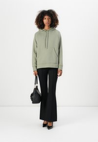 Olivengrøn hoodie med en kængurulomme, parret med sorte flagrende bukser. Modellen holder en sort håndtaske med guldhardware.